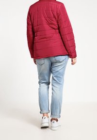 Red quilted jacket with stitched lines, blue jeans, and white sneakers. View from the back, showcasing the jacket's texture and length.