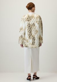 Vörös hajú nő kontyba fogva, bézs és barna mintás steppelt kabátban, fehér hosszú szoknyában és fekete lapos sarkú cipőben áll, háttal a falnak.