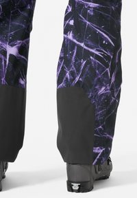 Sneeuwbroek met een zwart en paarse abstracte print, met versterkte zwarte onderste delen en bevestigde zwarte skischoenen.