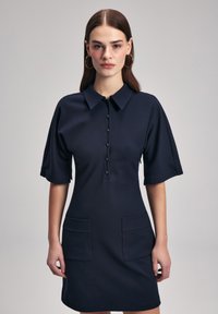 Navy blue lyhythihainen mekko, jossa on kaulus, etupainonappi, kaksi suurta jääkaappitaskua ja istuva malli. Sujuva rakenne.