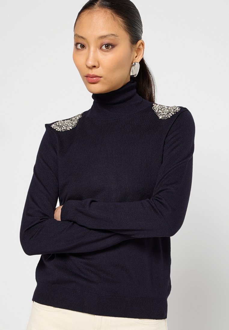 Donna con i capelli scuri raccolti in una bassa coda di cavallo, che indossa un maglione a collo alto blu navy con decorazioni gioiello sulle spalle e orecchini ovali argentati.