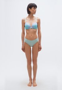 Γυναίκα όρθια, ξυπόλυτη, φοράει ανοιχτό μπλε εσώρουχο με ροζ δαντελένιες λουλουδένιες λεπτομέρειες, κοιτάζει προς τα δεξιά σε λευκό απλό φόντο.