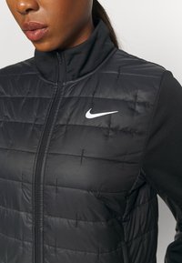 Czarna puchowa kamizelka o pikowanej teksturze, z wysokim kołnierzem i zapięciem na zamek. Posiada białe logo Nike na górnej lewej stronie.