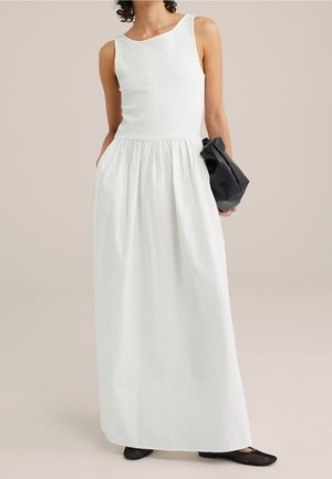 Vrouw draagt een witte mouwloze maxi-jurk met een geplooide taille, houdt een zwarte clutch vast en draagt zwarte mesh ballerina's.