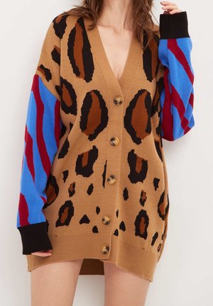 Kvinde iført en brun leopardmønstret cardigan med blå og røde stribede ærmer, knappet foran, hænder løftet nær skulderen.