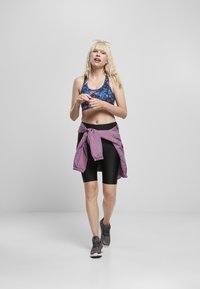 Svarte ribbede sykkelshorts, lilla vindjakke knyttet rundt livet, og en blåmønstret sports-BH. Grå joggesko fullfører den atletiske stilen.