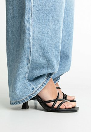 Pieds portant des sandales à talons noires à brides avec des bouts carrés, partiellement recouverts par un jean large en denim bleu clair.