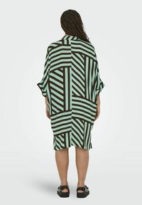 Sukienka w print w kolorze miętowej zieleni i ciemnobrązowym geometrycznym wzorze. Luźny krój, długość do kolan, z szerokimi rękawami i bocznymi rozcięciami.