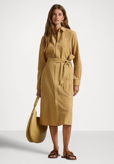 Kvinne iført en tanfarget kjole med knapper og belte knyttet i midjen, som holder en matchende tanfarget håndveske og har på brune sandaler med spenne.