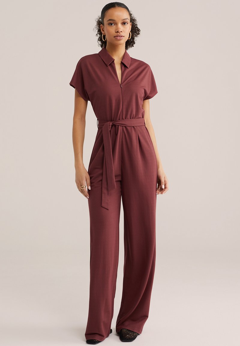 Naine, kes kannab lühikeste varrukatega pruuni kombekat krae, esiluku, vööga vöökohal ja laia säärega pükstega, seisab ühevärvilise tausta ees.