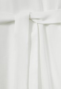 Tessuto bianco con una consistenza liscia, annodato a fiocco in vita. Caratteristiche di design semplici, che enfatizzano linee pulite e minimalismo.