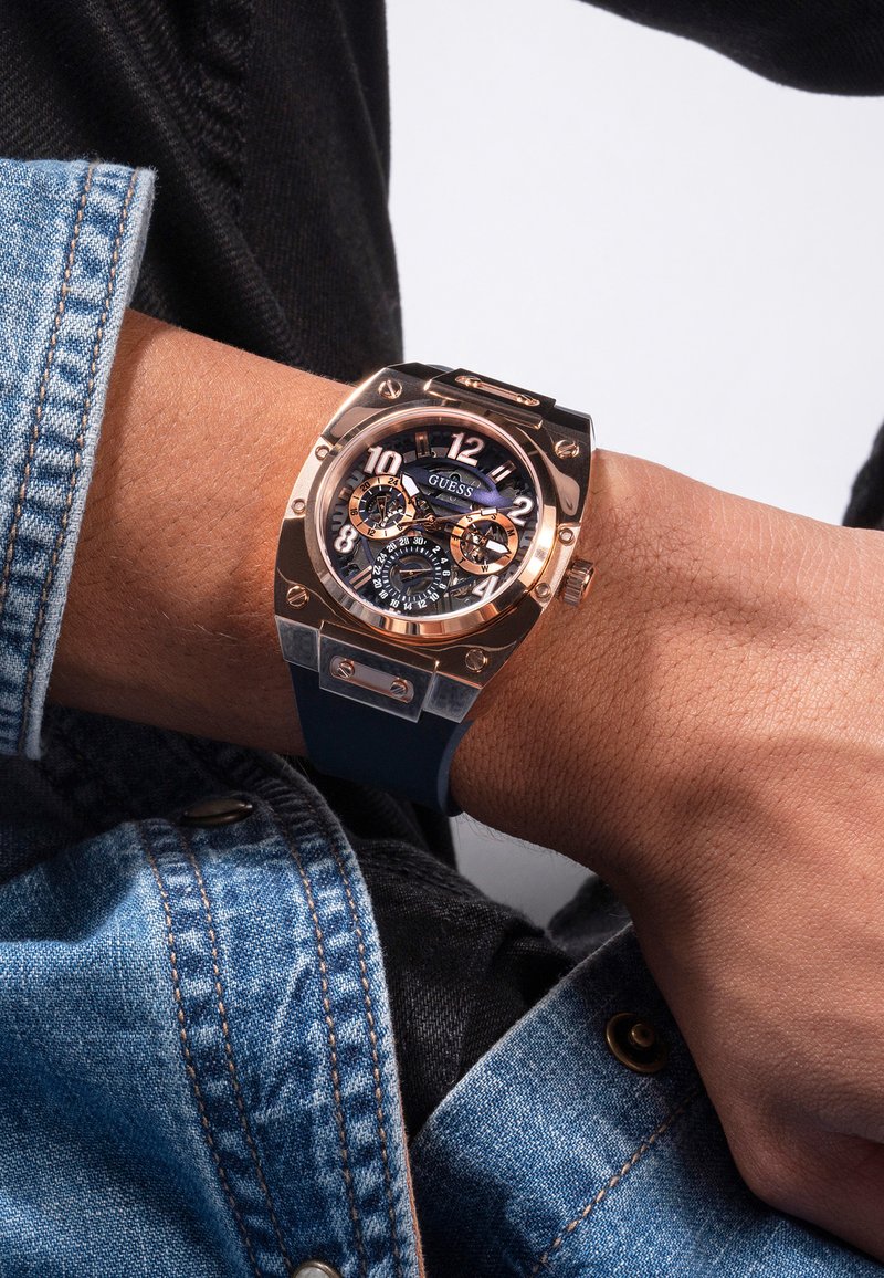 Sorte og rose-guld armbåndsur med en klar sag, store hvide tal og blå detaljer, båret på et håndled. Denimbetræk synligt.