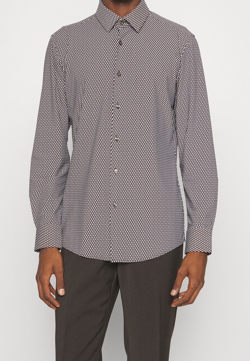 Homme portant une chemise ajustée à manches longues avec un petit motif losange noir et blanc et un pantalon sombre sur un fond uni.