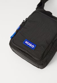 Svart stoffveske med en teksturert overflate. Har en blå "HUGO"-logo, glidelås og justerbar reim. Kompakt, rektangulært design.