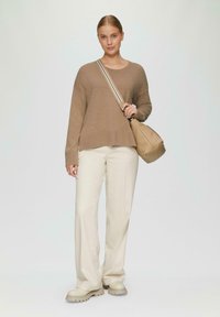 Frau steht und trägt einen beigen Pullover, cremefarbene weit geschnittene Hose, klobige beige Schuhe und eine beige Schultertasche mit gestreiftem Riemen vor einem weißen Hintergrund.