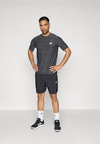 Musta urheilupaita, jossa on harmaa raidallinen kuvio, yhdistettynä mustiin shortseihin. Sisältää valkoisia logoelementtejä ja on yhdistetty mustiin urheilukenkiin sekä valkoisiin sukkiin.