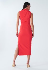 Sarkana bezroku kleita ar pieguļošu siluetu, sānu šķēlumu un gludu tekstūru. Aizmugurē ir mīksti krokojumi. Modelis valkā kleitu kopā ar ādas krāsas augstpapēžu kurpēm.