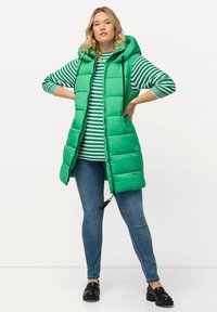 Grüne, plüschige Weste mit Kapuze, mit Reißverschluss und seitlichen Taschen, getragen über einem gestreiften grün-weißen Oberteil und blauen Jeans.