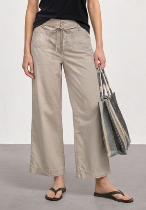 Frau trägt beige, weite Hosen und ein schwarzes Oberteil, hält eine gestreifte Tragetasche und trägt schwarze Flip-Flop-Sandalen.