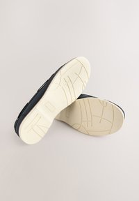 Chaussures noires à enfiler avec semelles en caoutchouc blanches présentant des motifs géométriques, exposées sur un fond clair.
