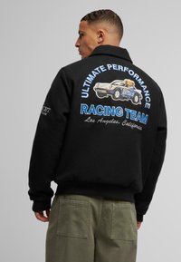 Black sweatshirt with a large racing graphic on the back, featuring a cartoon car and text in blue and white. Styled with loose olive pants.