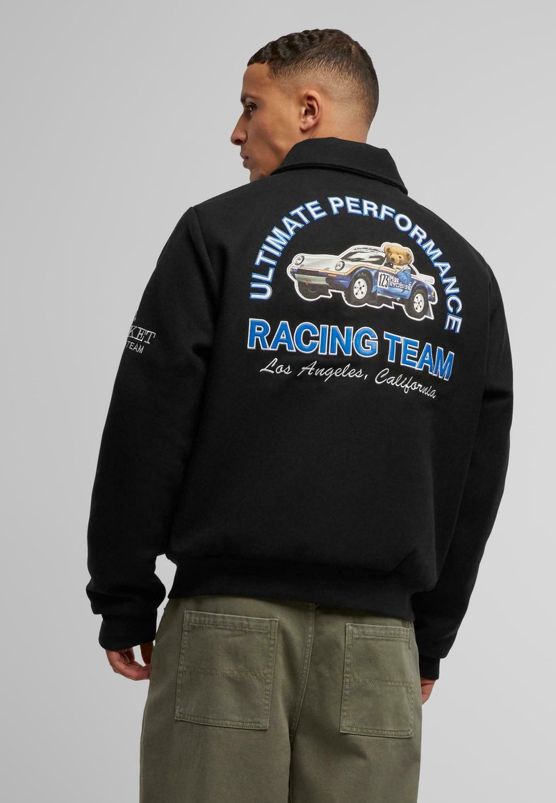 Black sweatshirt with a large racing graphic on the back, featuring a cartoon car and text in blue and white. Styled with loose olive pants.