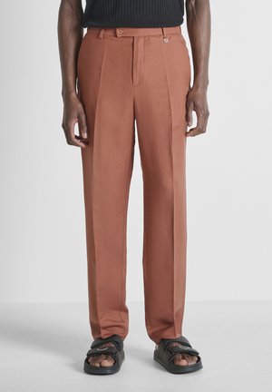 Homme portant un pantalon sur mesure couleur rouille et des sandales noires à bout ouvert, debout devant un fond clair uni.