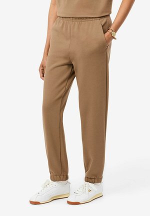 Person trägt braune Jogginghosen mit elastischem Bund und weiße Sneakers, steht mit einer Hand in der Tasche vor einem einfarbigen Hintergrund.