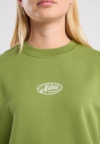 Groene sweatshirt van textuurstof met een ronde hals en een witte "Melrose" logo met decoratieve elementen.