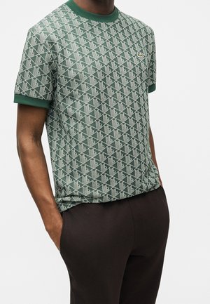 Homme portant une chemise à manches courtes verte à motif géométrique et un pantalon foncé, mains dans les poches, sur fond blanc uni.