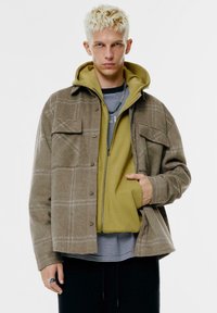 Ruutukuvioinen ruskea takki harmailla yksityiskohdilla vihreän hupparin päällä, musta-valkoisen raidallisen paidan päällä. Raskaampi kangas, jossa napit yksityiskohtana.