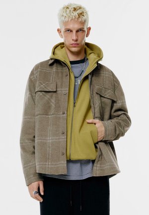 Bruine geruite jas met grijze accenten over een groene hoodie, gelaagd over een zwart-wit gestreept shirt. Getextureerde stof met knoopdetails.