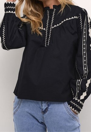 Blouse noire à manches longues avec des motifs géométriques blancs brodés sur la poitrine, les épaules et les manches, portée avec un jean bleu clair.