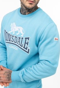 Világoskék pulóver puha anyagból, hímzett oroszlán logóval és merész "Lonsdale" felirattal az elején. Bordázott mandzsetta tartozik hozzá.