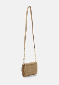 Malá béžová prošívaná crossbody taška se zlatými řetězovými ozdobami a tenkým koženým popruhem, zavěšená na bílém pozadí.