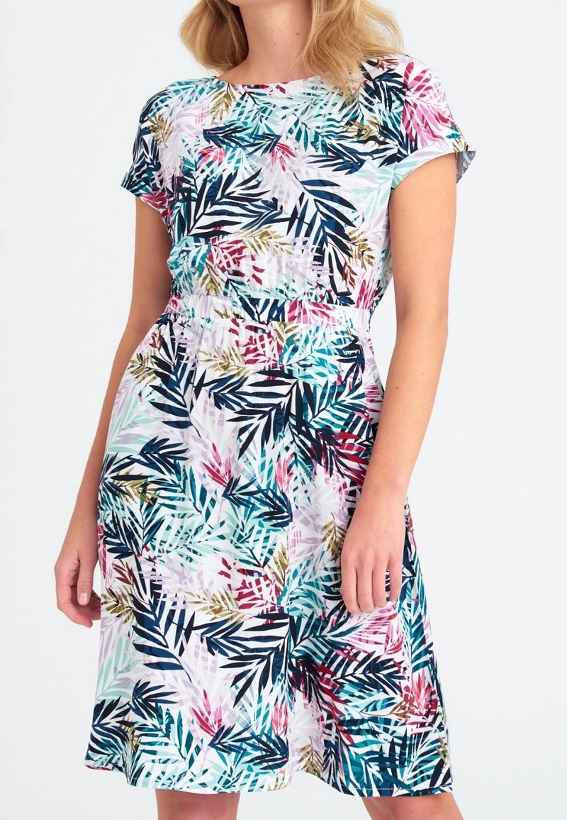 Femme portant une robe courte arrivant aux genoux, à manches courtes, avec un imprimé feuilles tropicales multicolores et une taille élastiquée.