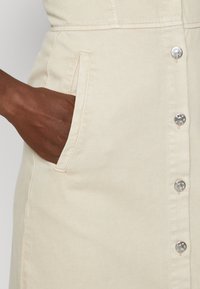 Bežinis audinio suknelė su mygtukų užsegimu, sidabriniais mygtukais ir šoninėmis kišenėmis. Tekstūra lygi, o dizainas atsitiktinis.