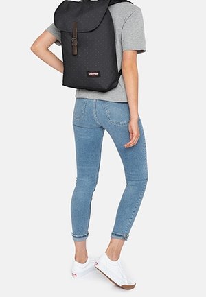 Černý batoh s kulatým klopovým uzávěrem, puntíkovaným vzorem a detaily hnědého koženého popruhu. Nošený na zádech, v kombinaci s přiléhavými modrými džínami a bílými teniskami.