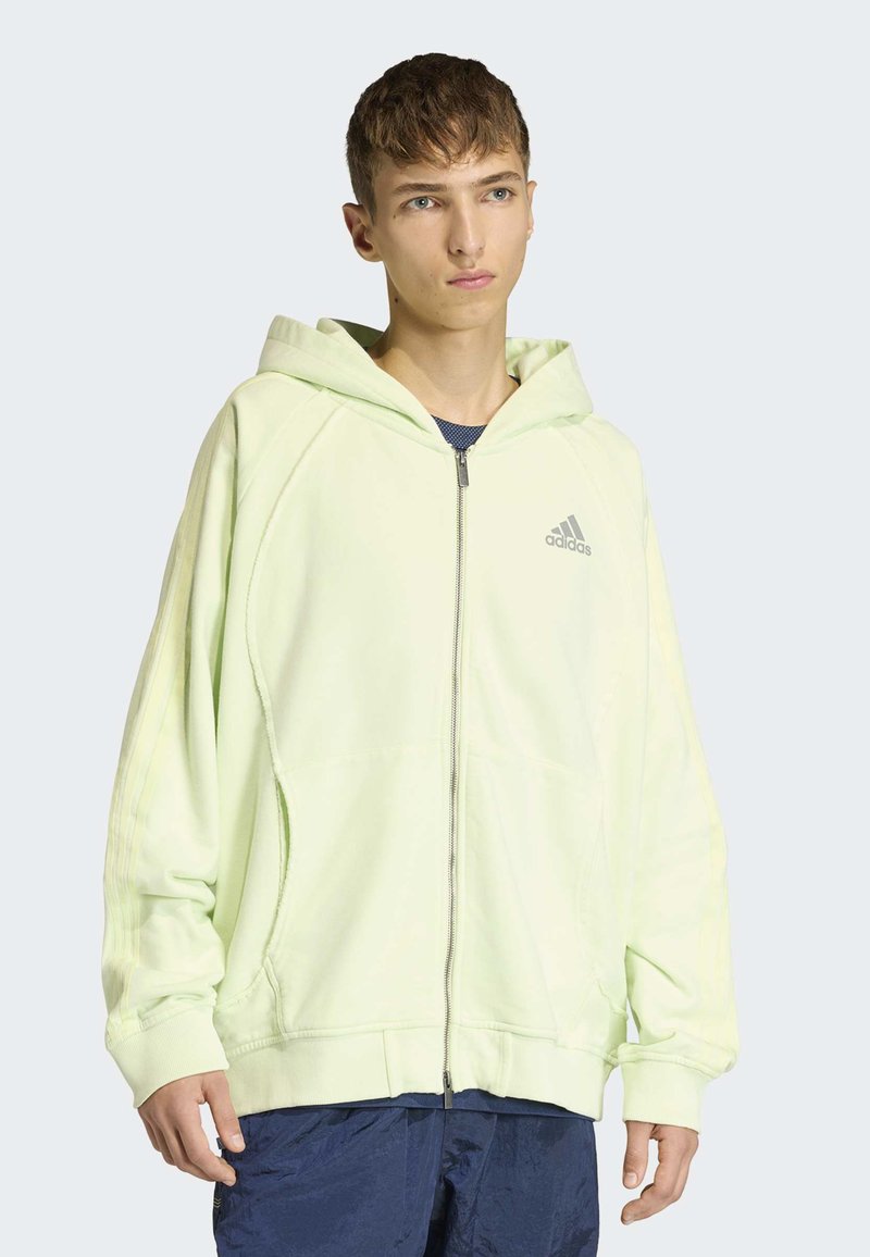 Sweat à capuche zippé vert clair avec une grande capuche, doté de poignets et d'un ourlet côtelés, et du logo Adidas sur la poitrine gauche. Porté par-dessus un haut bleu marine.