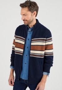 Cardigan en tricot bleu marine avec un col haut, ornée de rayures horizontales beiges, rouille et bleues. Deux poches latérales, poignets et ourlet côtelés.