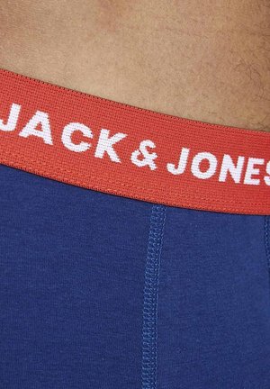 Nahaufnahme von blauer Unterwäsche mit rotem elastischem Bund und weißem "JACK & JONES" Schriftzug an der Taille einer Person.