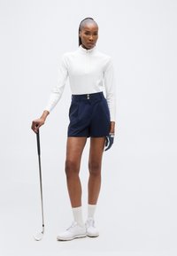 Γυναίκα με λευκή μακρυμάνικη μπλούζα, σκούρα μπλε σορτς, γάντι του γκολφ και λευκά παπούτσια κρατώντας ένα μπαστούνι γκολφ μπροστά σε λευκό φόντο.