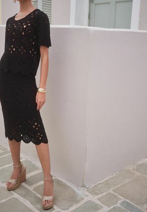 Mujer con conjunto de blusa y falda de encaje negro con bordes en forma de ondas, sandalias cuña color marrón claro y una pulsera dorada, de pie sobre pavimento de piedra.