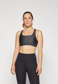 Mujer joven con el cabello recogido, vistiendo un sujetador deportivo negro y leggings de cintura alta, de pie contra un fondo claro y sencillo.