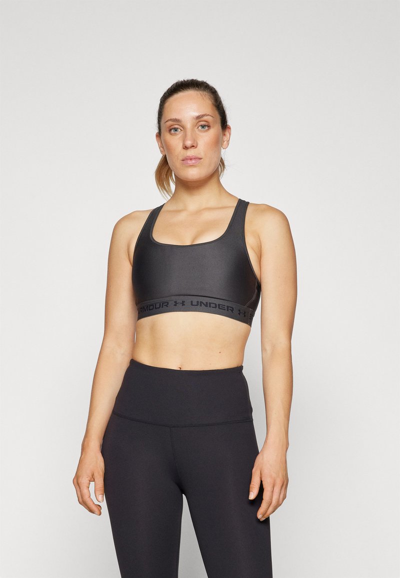 Mujer joven con el cabello recogido, vistiendo un sujetador deportivo negro y leggings de cintura alta, de pie contra un fondo claro y sencillo.