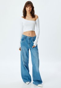 Witte lange mouwen crop top met een ribbeltextuur, gecombineerd met lichtblauwe wijd uitgesneden jeans. De outfit wordt aangevuld met witte puntige schoenen.