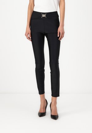 Pantaloni neri aderenti con una cintura larga che presenta un dettaglio metallico. Tessuto liscio, design sartoriale e gamba affusolata. Indossati con tacchi neri.