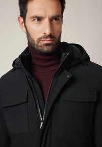 Czarna kurtka na zewnątrz z zamkiem z przodu, dwiema kieszeniami na piersi i kapturem, noszona na bordowym golfie; gładka tekstura materiału.