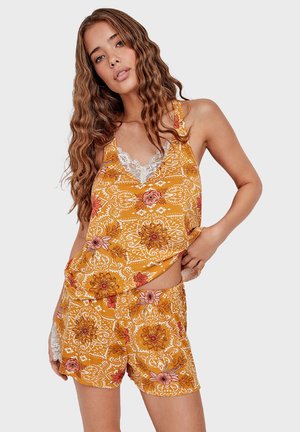 Woman with long wavy hair wearing mustard yellow floral pajama set with lace trim, posing with one hand on hip against plain background.