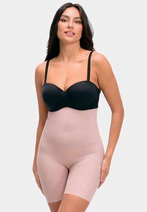 Femme aux longs cheveux foncés portant un soutien-gorge noir et un short gainant taille haute rose clair sur un fond clair uni.